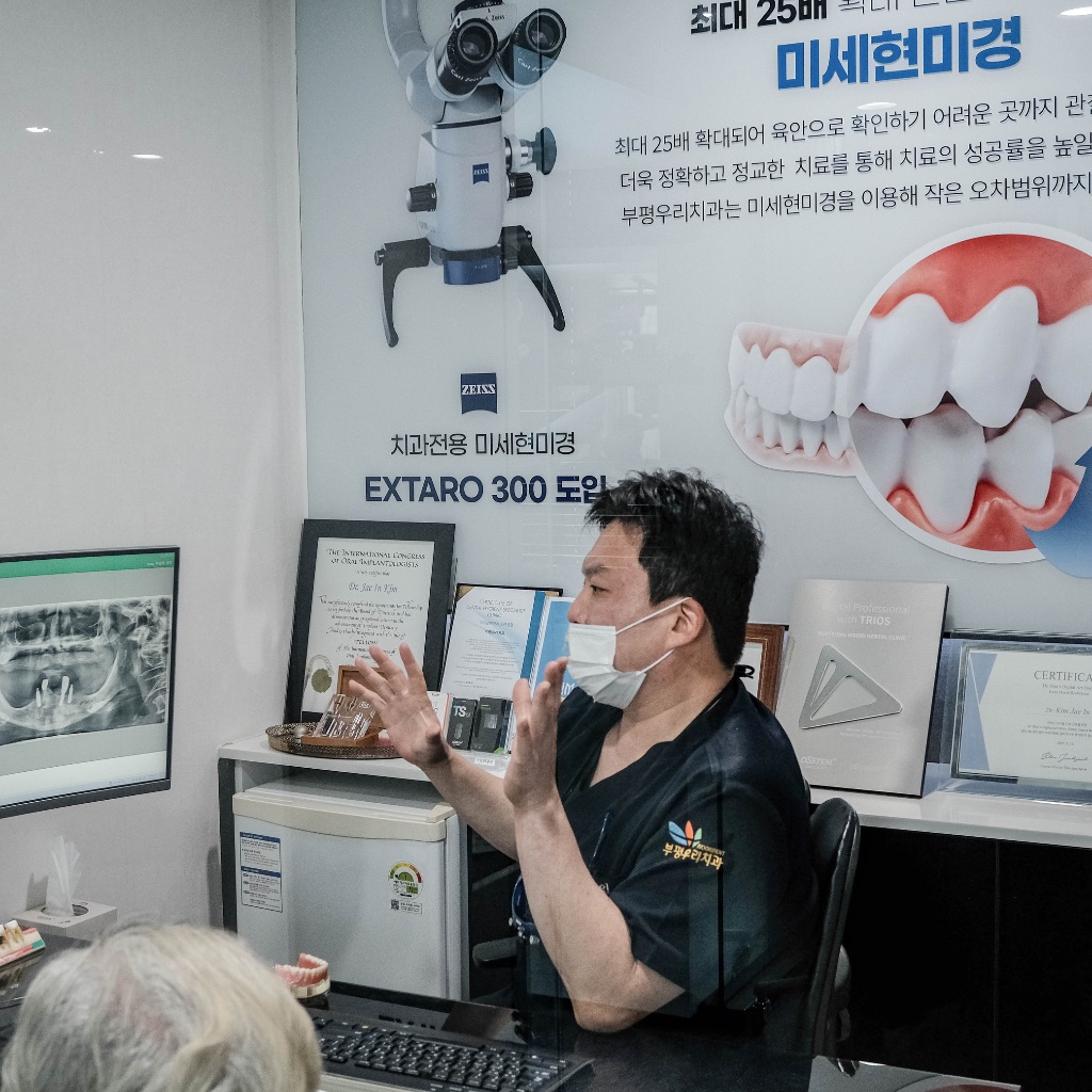 환자에게 직접 손짓으로 설명하는 김재인 대표원장 — 미세현미경 EXTARO 300 도입 안내 패널 앞